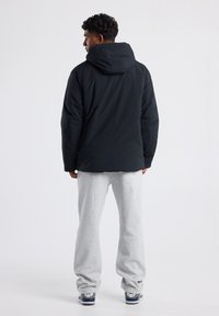 Chaqueta negra con capucha de textura lisa, llevada sobre pantalones de chándal grises. La chaqueta es de longitud media y presenta un diseño sencillo sin logotipos visibles.