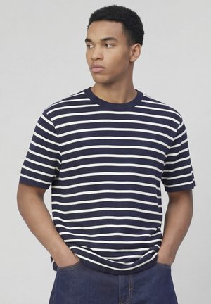 Jeune homme aux cheveux courts et bouclés portant une chemise à manches courtes rayée bleu marine et blanche, mains dans les poches, regardant à gauche.