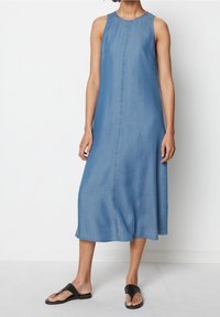 Vestido sin mangas de mezclilla en azul claro, con un corte recto, bolsillos laterales y costuras en contraste a lo largo del centro. Combinado con sandalias negras.