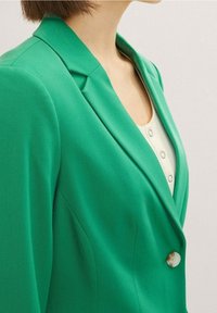 Femme portant un blazer vert vif sur un haut blanc boutonné, montrée de l'épaule à la moitié de la poitrine sur un fond neutre.
