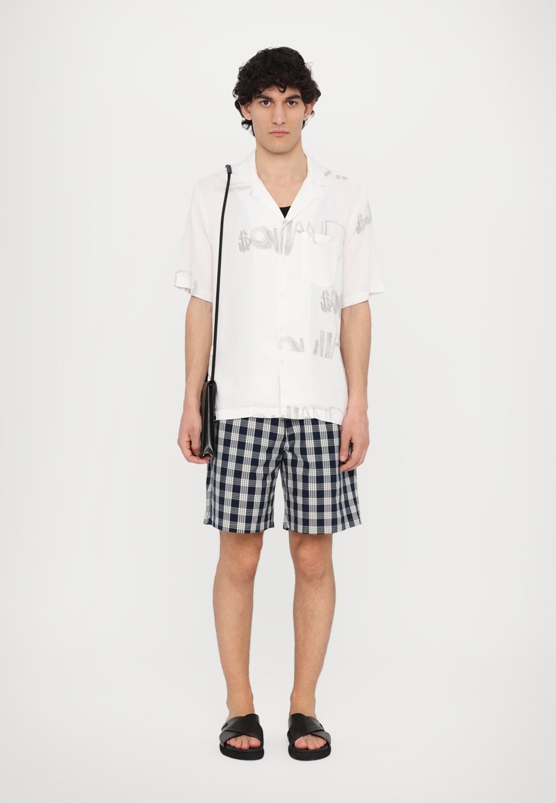 Chemise blanche à manches courtes avec des graphismes gris, débardeur noir, shorts à carreaux bleu marine et sandales noires. Le modèle tient un sac bandoulière noir.