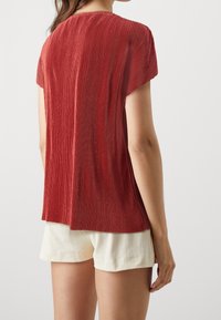 Top rojo con pliegues y mangas cortas, hecho de una tela texturizada, con un corte holgado y un dobladillo curvado, combinado con pantalones cortos color crema.