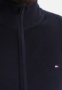 Námořnický svetr na zip z měkkého pleteného materiálu, s vysokým límcem a malou logovou aplikací v červené, bílé a modré barvě na hrudi.