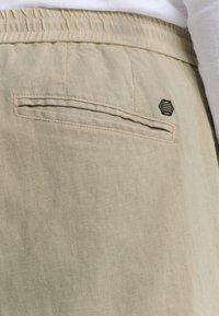 Beige linnen broek met een achterzak voorzien van een gestikt detail en een klein hexagonaal logo accent. Zachte textuur, casual ontwerp.