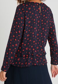 Blusa navy con motivo floreale rosso, maniche lunghe e orlo a peplum. Presenta una chiusura a fessura sul retro e una texture di tessuto liscia.