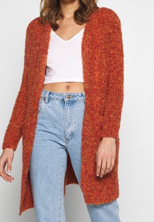 Pitkä, avonainen etupuoli kardigan, valmistettu teksturoidusta oranssista materiaalista, päällä valkoinen toppi. Yhdistetty vaaleansinihin korkeavyötäröisiin farkkuihin.