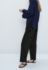 Dunkelblauer Strickpullover mit weiten Ärmeln, kombiniert mit schwarzen Cargo-Hosen mit seitlichen Taschen, ergänzt durch flache schwarze Sandalen.