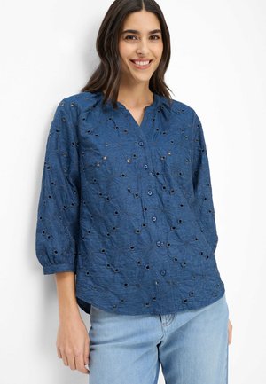 Frau lächelt, trägt ein blaues Hemd mit Knöpfen und augenförmigen Blumenmustern sowie hellblaue Jeans, steht vor einem weißen Hintergrund.