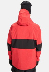 Rote wasserdichte Jacke mit einem schwarzen horizontalen Streifen, mit Kapuze, Gittermuster und elastischen Bündchen. Rückansicht zeigt glatte Struktur.