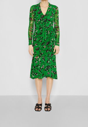 Grøn midi-kjole med sort og hvidt blomsterprint, lange gennemsigtige ærmer, V-udskæring og sorte sandaler med hæl og remme på en ensfarvet baggrund.