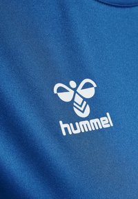 Kék sportmez sima, szintetikus anyagból, fehér logóval, stilizált formákkal és az "hummel" márkanevével az alján.