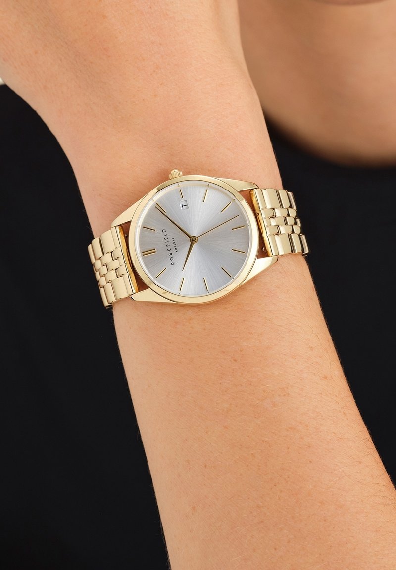 Montre-bracelet en or avec un cadran argenté et un design minimaliste portée au poignet d'une personne indiquant l'heure 7h24.