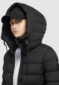 Zwarte pufferjacket met hoge capuchon, ritssluiting en verstelbare trekkoord. Bevat zichtbare stiksels en een logo-opdruk op de mouw.