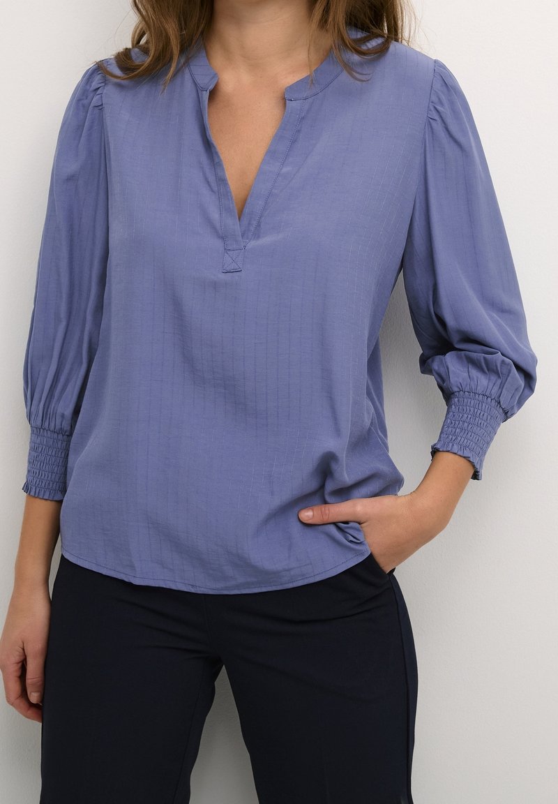 Blouse bleu clair avec un décolleté en V et des manches trois-quarts froncées. Elle présente un motif de rayures verticales subtiles et une coupe décontractée.
