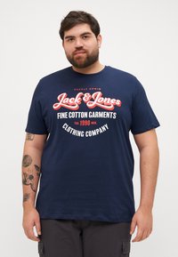 Tumesinine puuvillane t-särk, millel on valge ja punane "Jack & Jones" logo, lõdva lõike ja lühikeste varrukatega. Esitletud mudelil.