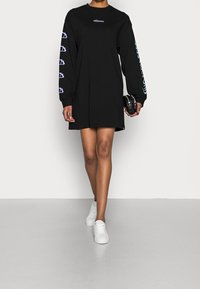 Robe noire à manches longues avec logo sur la poitrine, imprimés graphiques sur les manches et une coupe ample. Portée avec des baskets blanches sur un fond neutre.