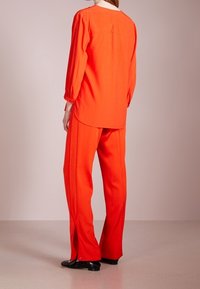 Czerwona koszula z długim rękawem i zaokrąglonym dołem, zestawiona z dopasowanymi, luźnymi spodniami z bocznymi rozcięciami, wykonanymi z gładkiego materiału.