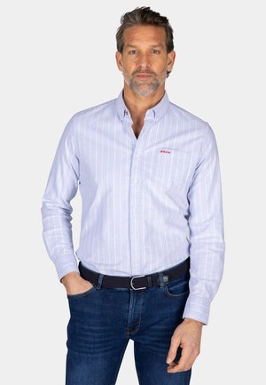 Lichtblauwe button-up shirt met witte verticale strepen, borstzak en rood logo. Draagt bij donkere blauwe jeans en een gestructureerde zwarte riem.