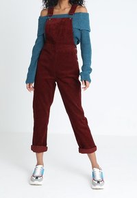 Person som bär uppvikta vinröda kordflorsbyxor, en off-the-shoulder turkos stickad tröja och vita sneakers med blåa och röda detaljer.