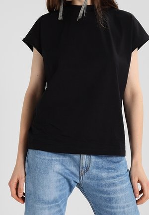 Woman wearing a plain black short-sleeve top and light blue denim jeans, accessorized with long silver tassel earrings.