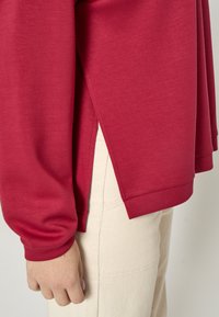 Punane pikemate varrukatega kampsun küljevõlvidel. Mugav tekstuur ja ümar ääreosa. Sobitatud helebeežide, kõrge vöökohaga pükstega, millel on nähtav õmblus.