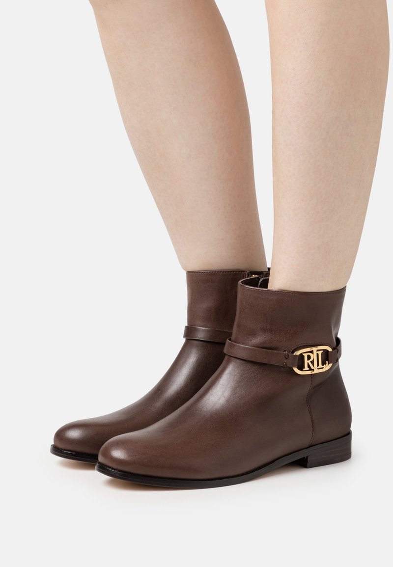 Bottes basses en cuir marron avec talons bas, fermeture éclair latérale et boucle dorée "RL" sur les sangles, portées sur des jambes nues.