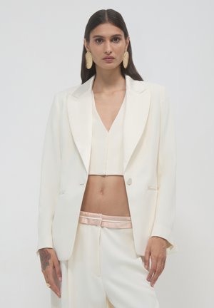 White tailored blazer with sharp lapels, paired with a matching cropped top and cream trousers featuring a pink satin waistband. Gold earrings.