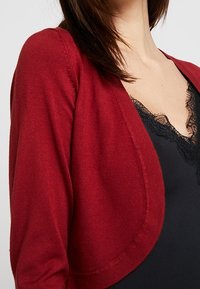 Röd stickad cardigan med en mjuk textur och öppen framsidan, bärs över en svart topp med spetsdetaljer vid halsringningen.