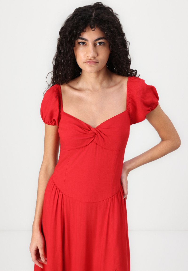 Rotes kurzes Kleid mit kurzen Ärmeln, herzförmigem Ausschnitt, verdrehtem Taillendetail und einem fließenden Rock. Hergestellt aus leichtem, glattem Stoff.