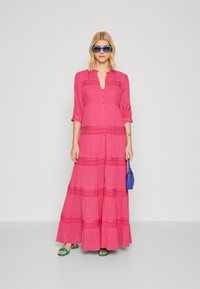 Vestido maxi rosa com botões na frente, mangas três quartos e detalhes de padrão horizontal. Usado com sandálias verdes e uma bolsa azul pequena.