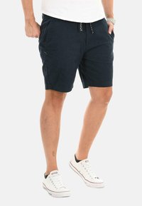 Navy blue cotton shorts with an elastic waistband, drawstring closure, two side pockets, and a relaxed fit. White sneakers complete the look.