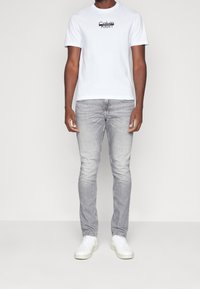 Camiseta de algodón blanca con logo negro en el pecho, combinada con jeans ajustados gris claro y zapatillas blancas, de pie sobre un fondo neutro.