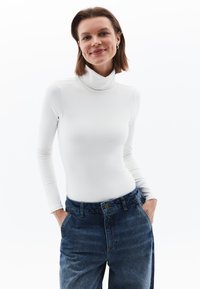 Weißes Turtleneck-Oberteil mit langen Ärmeln, figurbetontes Design, kombiniert mit hochgeschnittenen blauen Jeans aus Denim mit klassischem Knopfverschluss.