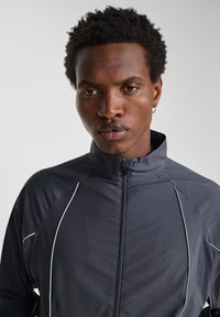 Grå zip-jacka med hög krage, med kontrasterande vit söm, en strukturerad tygyta och en figurnära design.