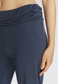 Marineblauwe leggings met een gedrapeerde tailleband, een gladde textuur en een aansluitende pasvorm, met een hand die rust op de zijkant.
