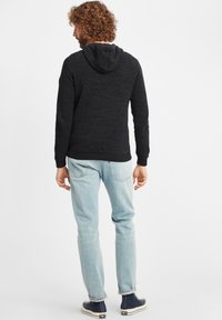 Schwarzer Strickhoodie mit strukturierter Oberfläche, gerippten Bündchen und Saum, kombiniert mit hellblauen Jeans und dunkelblauen Sneakers. Die Kapuze ist hinten sichtbar.