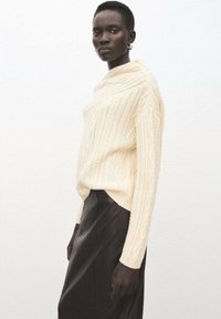 Kremfarvet kabelstrikket sweater med høj hals, kombineret med en mørk lædernederdel. Outfittet har lange ærmer og teksturerede mønstre.