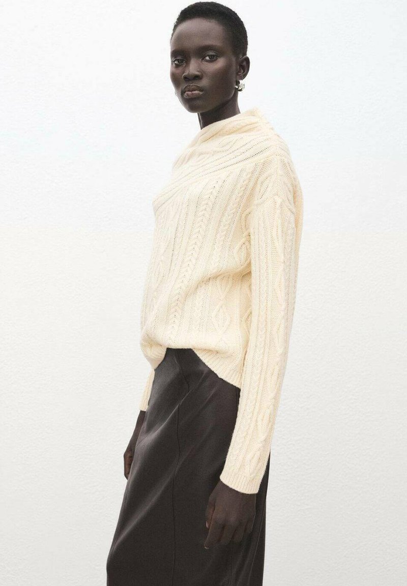 Kremfarvet kabelstrikket sweater med høj hals, kombineret med en mørk lædernederdel. Outfittet har lange ærmer og teksturerede mønstre.