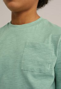 Krátký rukáv, mintově zelené tričko vyrobené z měkké tkaniny, s přišitými kapsičkami a malým vyšitým "R" na hrudi.