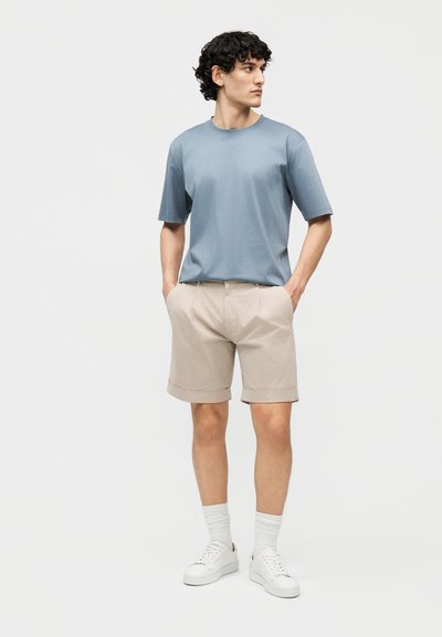 Jeune homme aux cheveux bouclés debout, les mains dans les poches, portant un t-shirt bleu à manches courtes, un short beige, des chaussettes blanches et des baskets blanches.