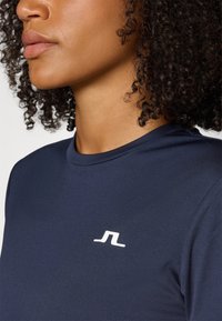 Camisola desportiva azul-marinho com decote redondo, mangas curtas e um detalhe de logó tipo branco no peito. Textura de tecido suave.