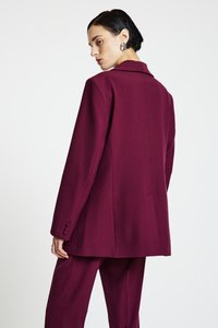 Blazer bordeaux sur mesure avec col à revers et poignets à un bouton. Tissu lisse avec une coupe structurée, mettant en valeur une fente au dos.