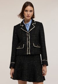 Donna che indossa un completo gonna nera texturizzata con dettagli bianchi e una camicia colletto azzurro chiaro, in piedi su uno sfondo neutro.