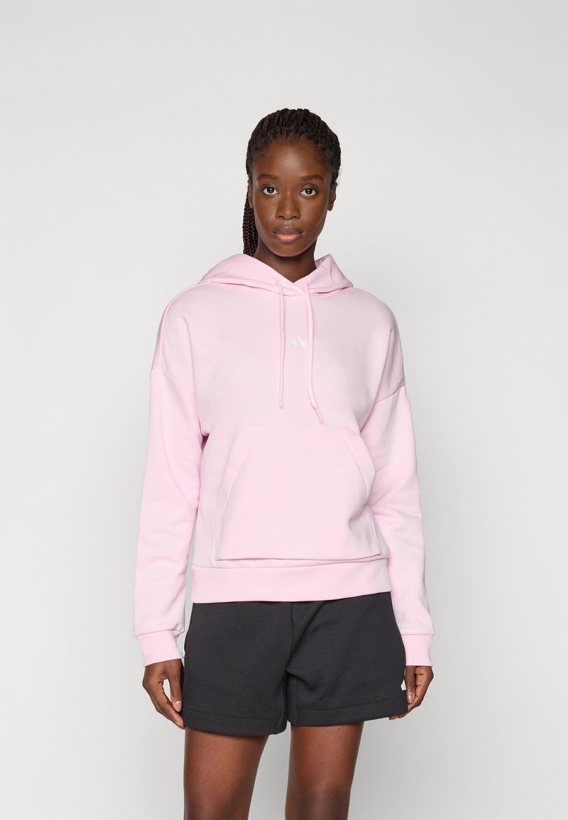 Sweat à capuche rose clair avec une poche avant, un cordon de serrage et un logo blanc. Associé à un short gris foncé. Tissu en coton, coupe décontractée.
