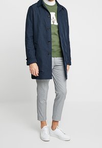 Námořnický venkovní kabát s předním zapínáním na knoflíky, nošený přes zelenou mikinu. Světle šedé kostkované kalhoty a bílé tenisky dokončují vzhled.