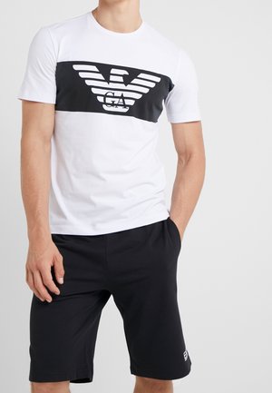 Hombre con una camiseta blanca con un gran logo negro y pantalones cortos negros, de pie con una mano en el bolsillo contra un fondo liso.