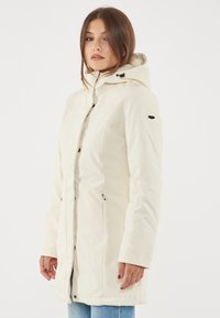 Giacca impermeabile beige chiaro, lunga fino al ginocchio, con cappuccio, zip frontale, due tasche laterali e dettagli in metallo nero. Tessuto liscio.