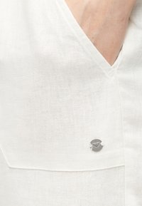 Prim-plan cu o haină dintr-un material alb texturat, cu un decolteu adânc în V și un mic buton rotund argintiu cu logo atașat lângă cusătură.