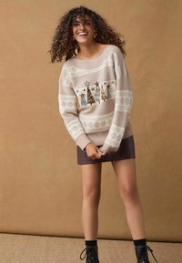 Maglione lavorato a maglia beige con motivi di fiocchi di neve bianchi e alberi ricamati con paillettes; abbinato a una mini gonna viola scuro.