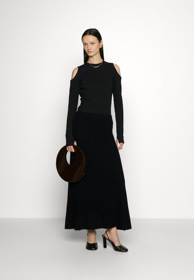 Schwarzer Cold-Shoulder-Pullover kombiniert mit einem gerippten schwarzen Maxirock. Das Model hält eine dunkelbraune, runde Handtasche und trägt schwarze High Heels.